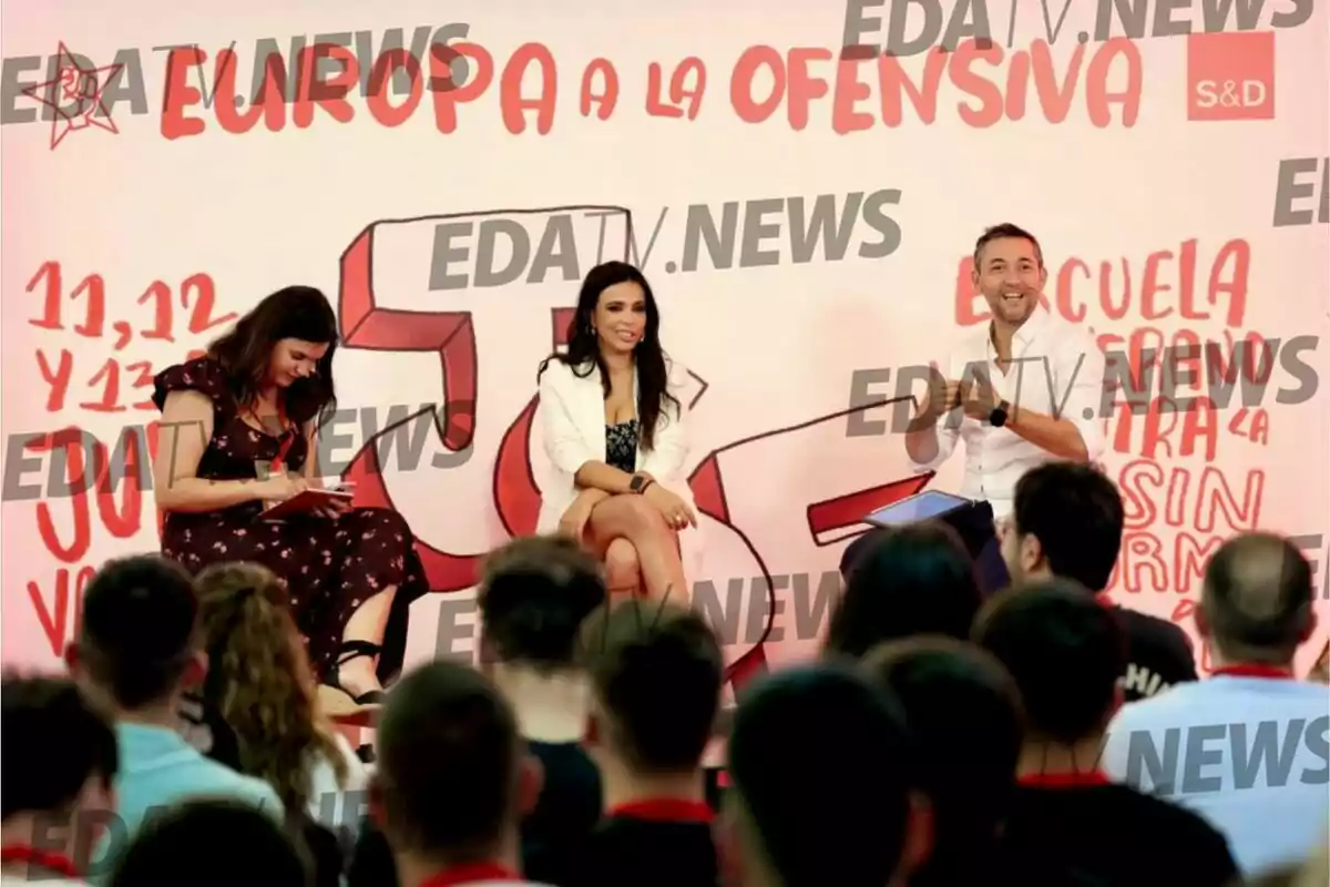 Tres personas sentadas en un escenario frente a una audiencia con un fondo que tiene letras rojas y decoraciones relacionadas con un evento europeo Tres personas sentadas en un escenario frente a una audiencia con un fondo que tiene letras rojas y decoraciones relacionadas con un evento europeo