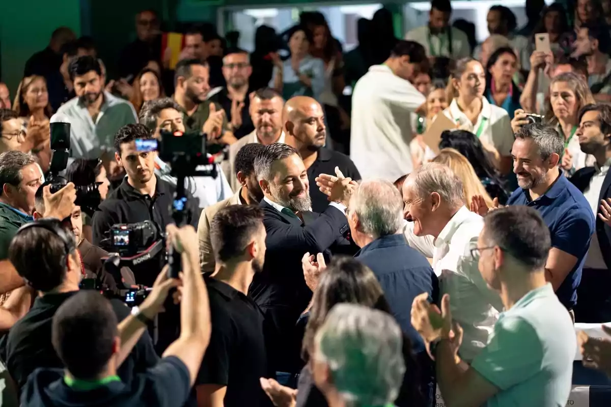 Un hombre con barba saluda sonriente a otra persona en medio de una multitud que aplaude y toma fotos en un evento concurrido.