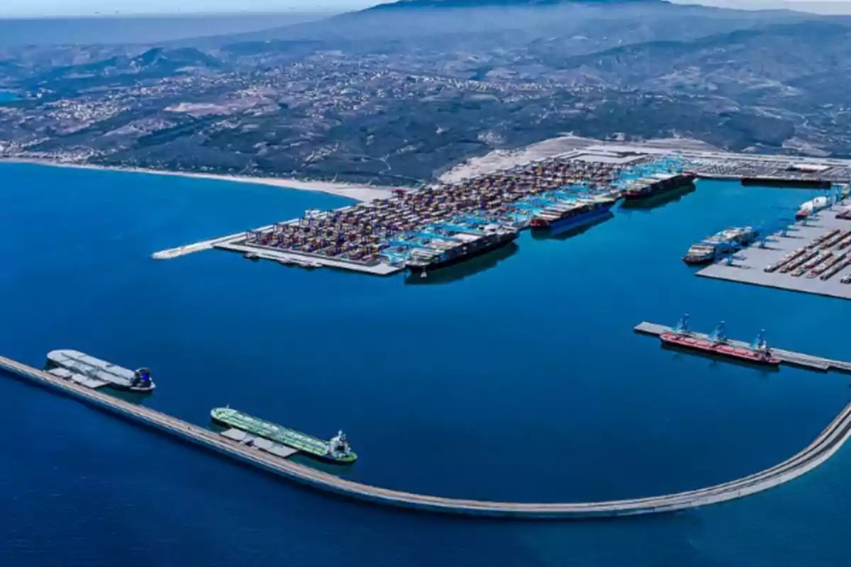 Vista aérea de un puerto marítimo con varios barcos y contenedores de carga junto a una ciudad y montañas al fondo Vista aérea de un puerto marítimo con varios barcos y contenedores de carga junto a una ciudad y montañas al fondo