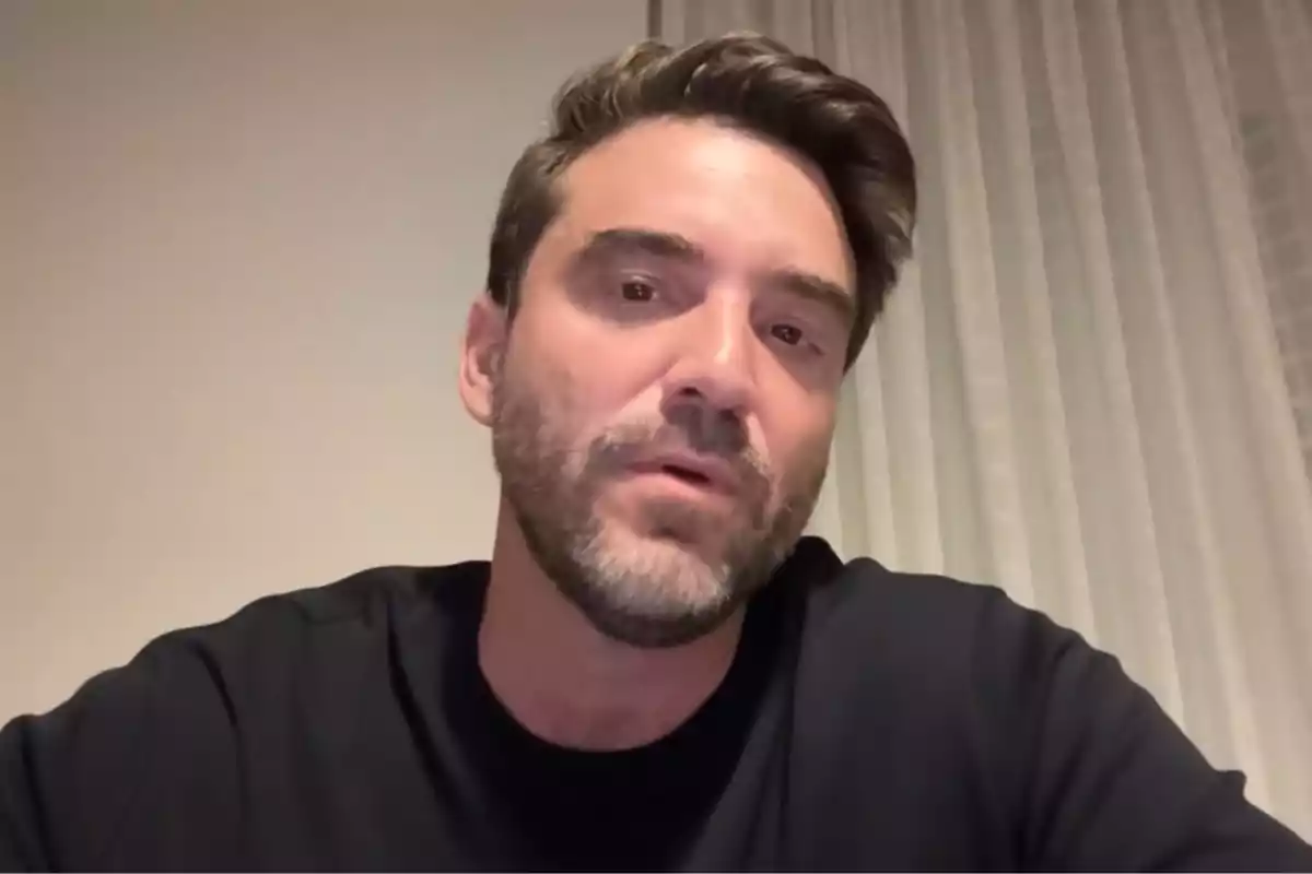 Hombre de cabello corto y barba recortada con camiseta oscura sentado frente a una cortina blanca Hombre de cabello corto y barba recortada con camiseta oscura sentado frente a una cortina blanca