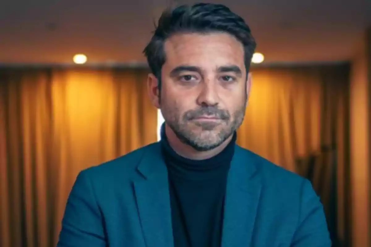 Hombre de cabello corto y barba con expresión seria vistiendo un saco azul y suéter negro frente a un fondo con cortinas y luces cálidas. Hombre de cabello corto y barba con expresión seria vistiendo un saco azul y suéter negro frente a un fondo con cortinas y luces cálidas.
