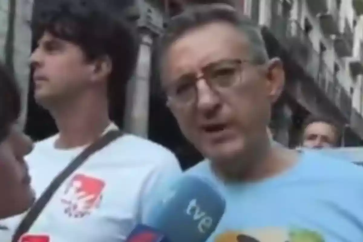 Un hombre con gafas y camiseta azul está siendo entrevistado en la calle por un medio de comunicación mientras otras personas aparecen al fondo. Un hombre con gafas y camiseta azul está siendo entrevistado en la calle por un medio de comunicación mientras otras personas aparecen al fondo.