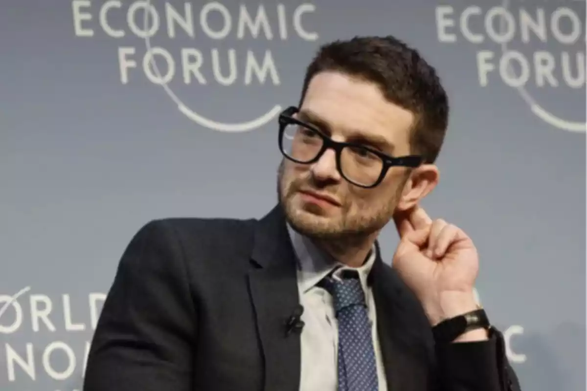 Hombre con gafas y traje sentado en un panel del Foro Económico Mundial tocándose la oreja Hombre con gafas y traje sentado en un panel del Foro Económico Mundial tocándose la oreja
