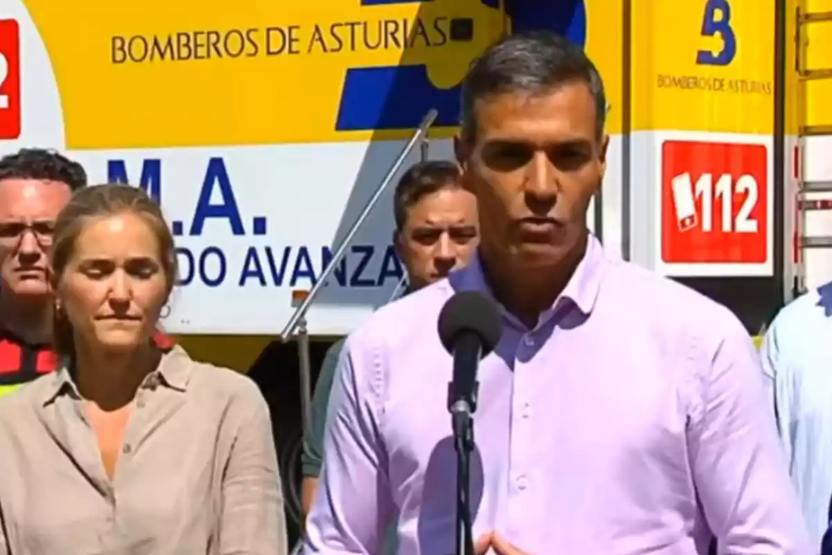 Un hombre con camisa clara habla frente a un micrófono acompañado de varias personas y un camión de bomberos de Asturias al fondo