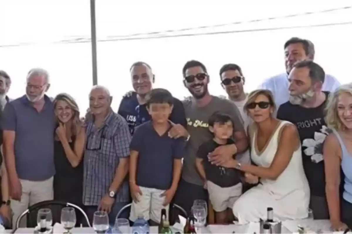 Un grupo de personas de diferentes edades posando juntas y sonriendo en una reunión social alrededor de una mesa con copas y botellas. Un grupo de personas de diferentes edades posando juntas y sonriendo en una reunión social alrededor de una mesa con copas y botellas.