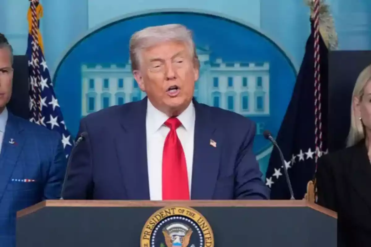 Hombre de cabello canoso con traje azul y corbata roja hablando en un podio presidencial con el escudo de Estados Unidos y banderas al fondo Hombre de cabello canoso con traje azul y corbata roja hablando en un podio presidencial con el escudo de Estados Unidos y banderas al fondo