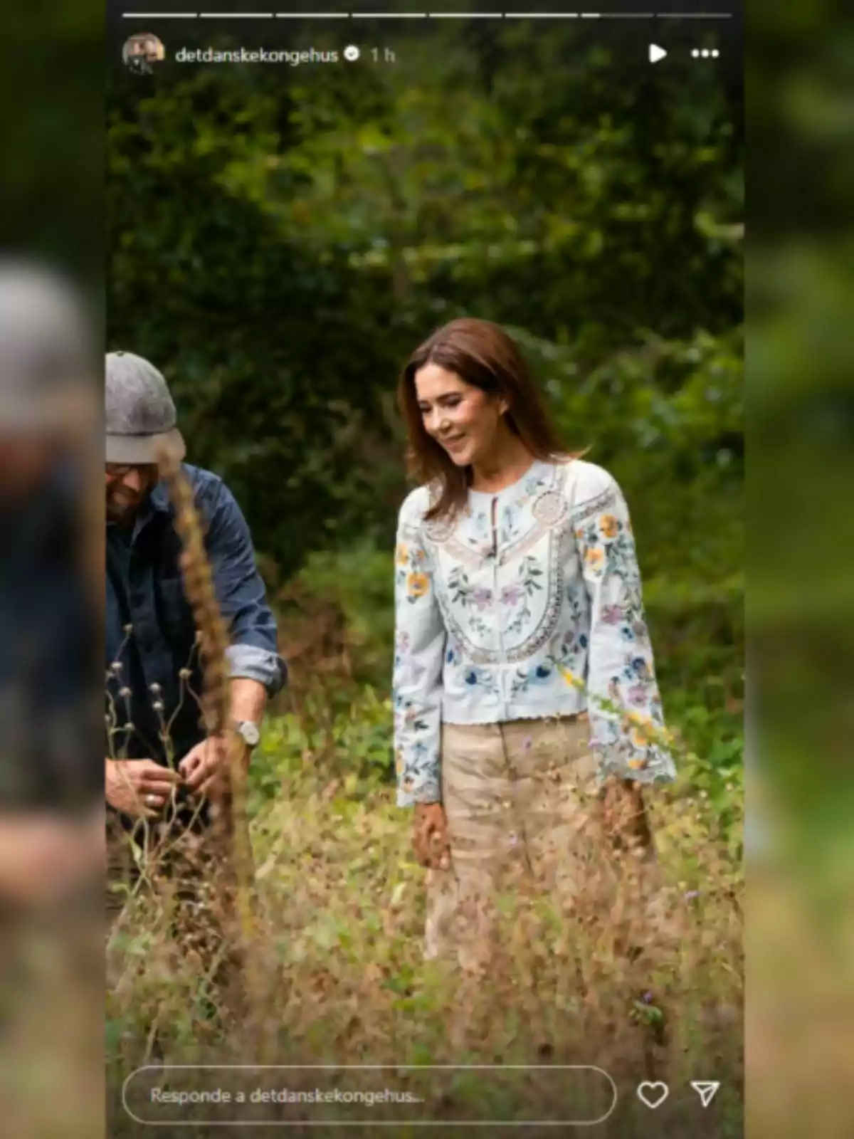 Mary of Denmark, smiling in an embroidered blouse and beige skirt, walking through a field surrounded by greenery next to a partially visible man Mary of Denmark, smiling in an embroidered blouse and beige skirt, walking through a field surrounded by greenery next to a partially visible man