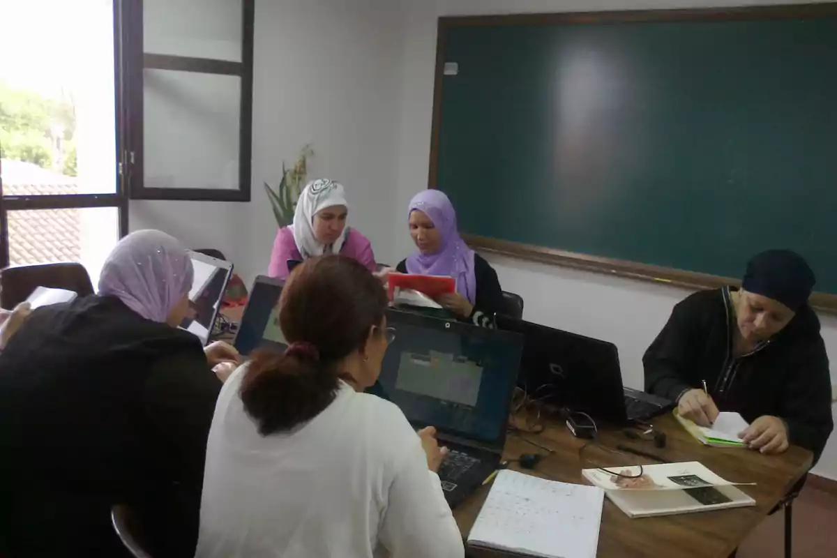 Cinco mujeres trabajan en una sala de clases usando computadoras portátiles y cuadernos, sentadas alrededor de una mesa frente a un pizarrón verde. Cinco mujeres trabajan en una sala de clases usando computadoras portátiles y cuadernos, sentadas alrededor de una mesa frente a un pizarrón verde.