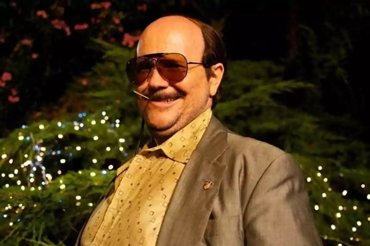 Hombre calvo con gafas de sol grandes, bigote y sonrisa, usando saco gris y camisa beige con estampado, fondo de luces y vegetación Hombre calvo con gafas de sol grandes, bigote y sonrisa, usando saco gris y camisa beige con estampado, fondo de luces y vegetación
