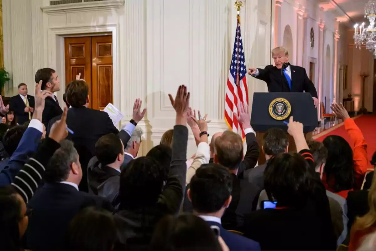 Una mujer hablando en un podio con el sello del Vicepresidente de los Estados Unidos mientras un hombre la observa en el fondo. Una mujer hablando en un podio con el sello del Vicepresidente de los Estados Unidos mientras un hombre la observa en el fondo.