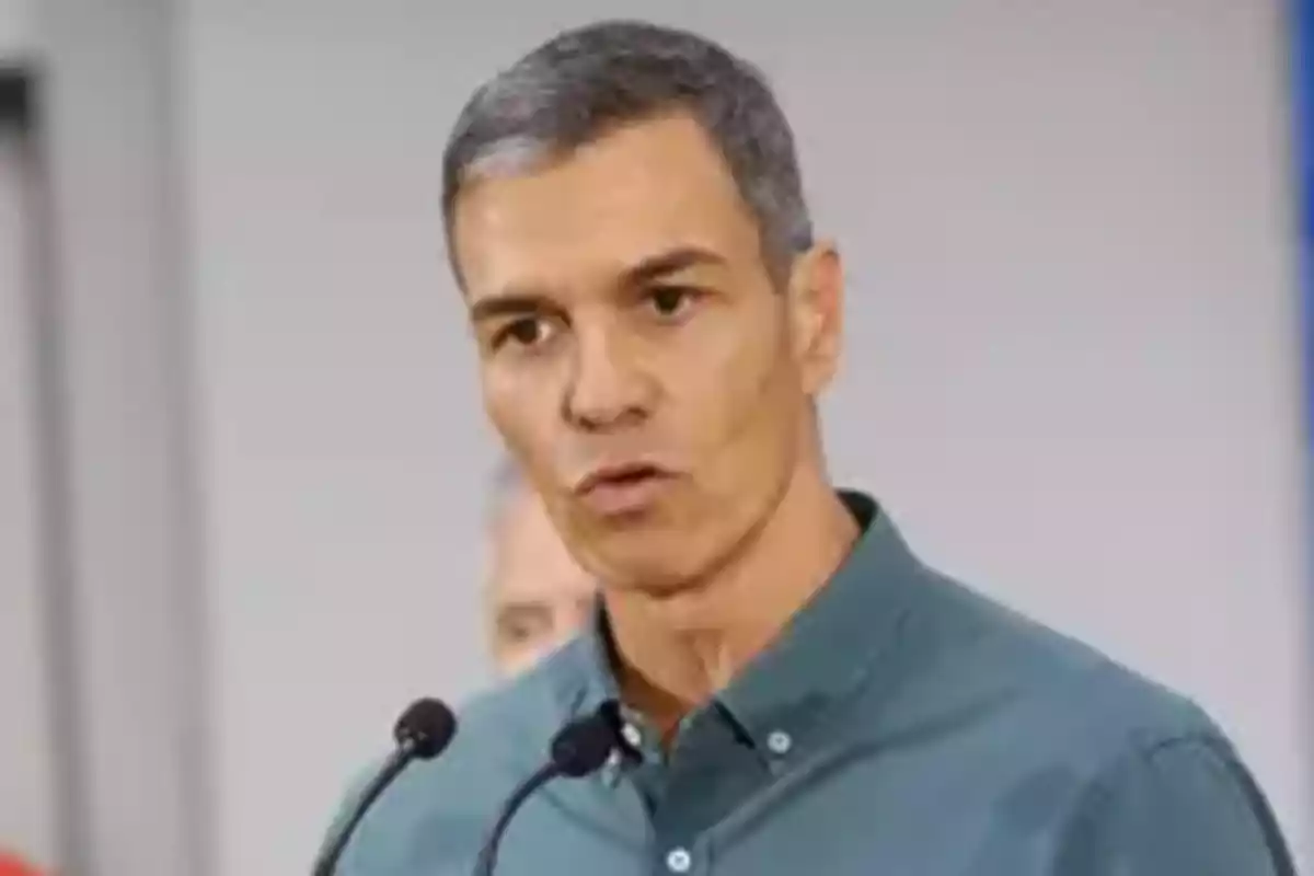 Hombre de cabello corto y canoso hablando frente a dos micrófonos con camisa verde Hombre de cabello corto y canoso hablando frente a dos micrófonos con camisa verde