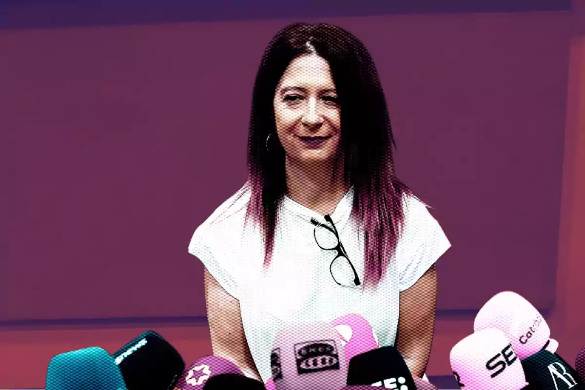 Mujer de cabello largo y oscuro con gafas colgadas en la camiseta blanca frente a varios micrófonos de colores en una rueda de prensa sobre fondo morado