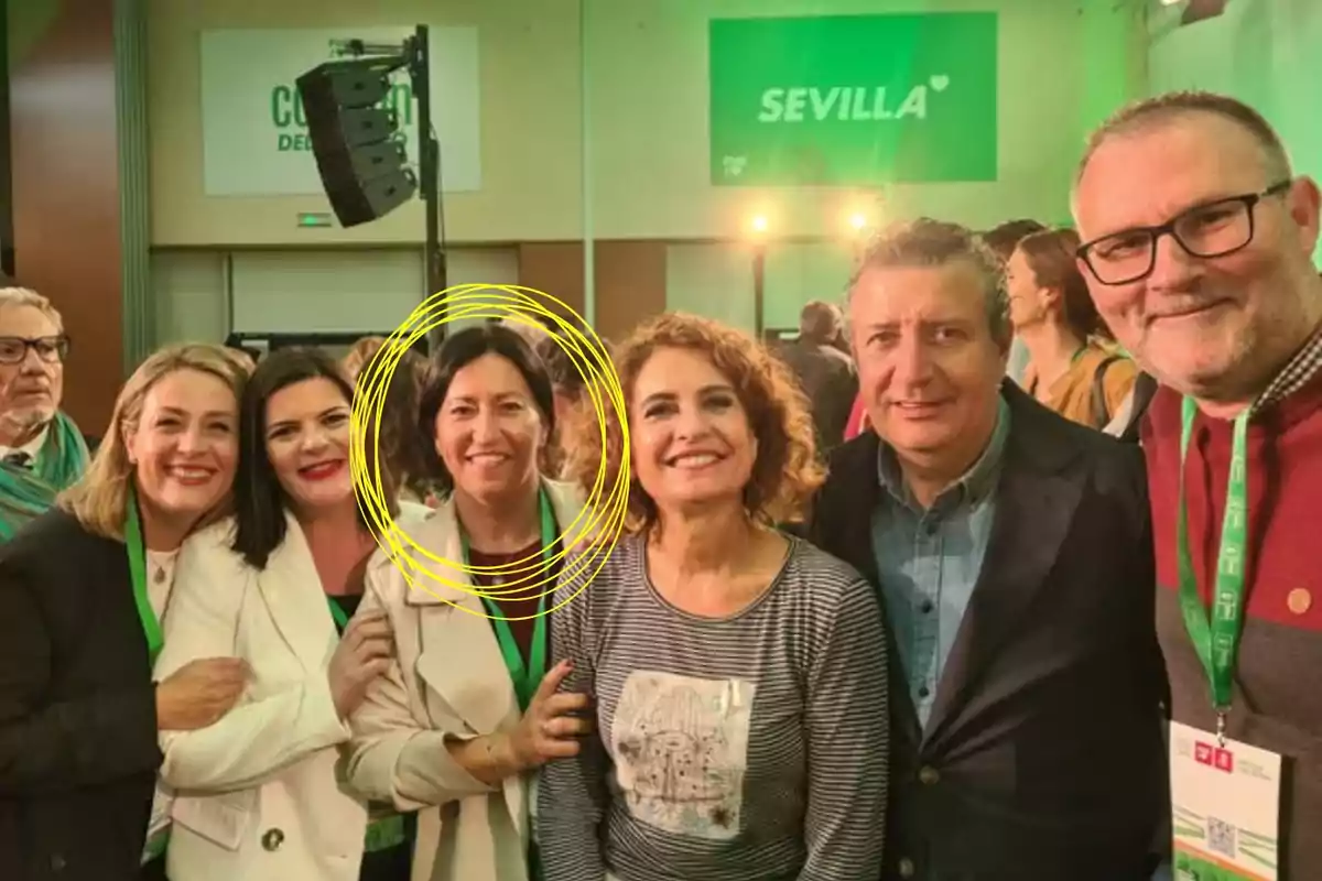 Grupo de personas posando y sonriendo en un evento con fondo verde y la palabra Sevilla, una mujer en el centro está resaltada con un círculo amarillo dibujado.