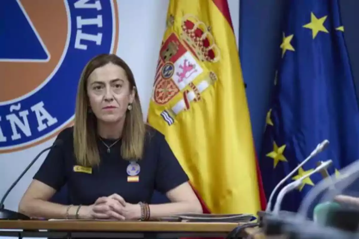 Mujer sentada en una mesa frente a micrófonos con banderas de España y la Unión Europea al fondo y un cartel de protección civil