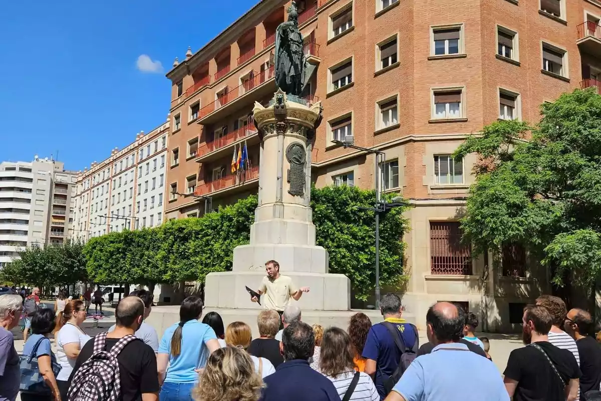 Un grupo de personas escucha a un guía turístico frente a una estatua en una plaza rodeada de edificios de ladrillo bajo un cielo despejado.