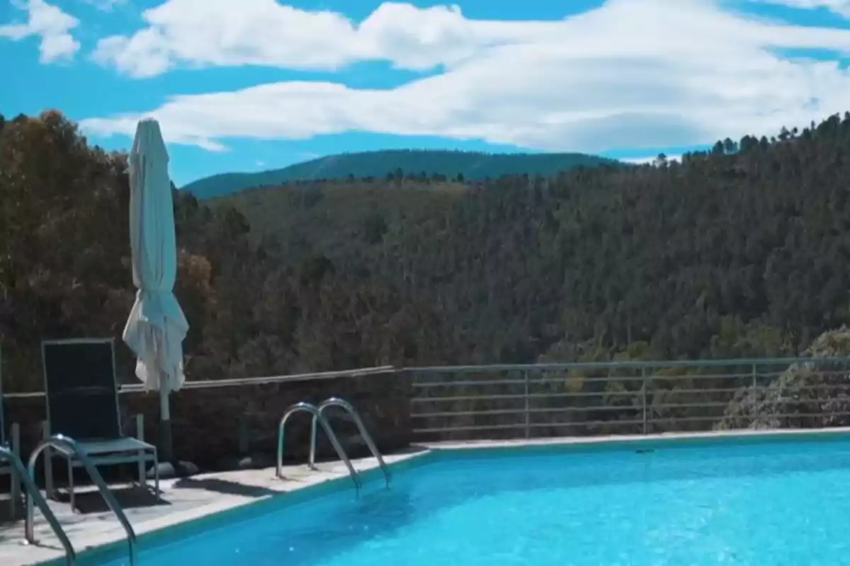 Piscina al aire libre con sillas y sombrilla cerrada, rodeada de montañas y árboles bajo un cielo azul con nubes Piscina al aire libre con sillas y sombrilla cerrada, rodeada de montañas y árboles bajo un cielo azul con nubes