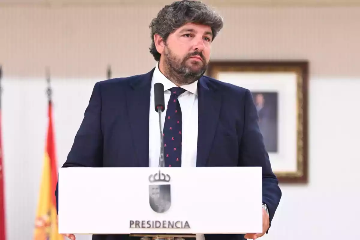 Hombre de cabello y barba oscuros con traje azul marino hablando en un atril de presidencia con banderas y un cuadro al fondo