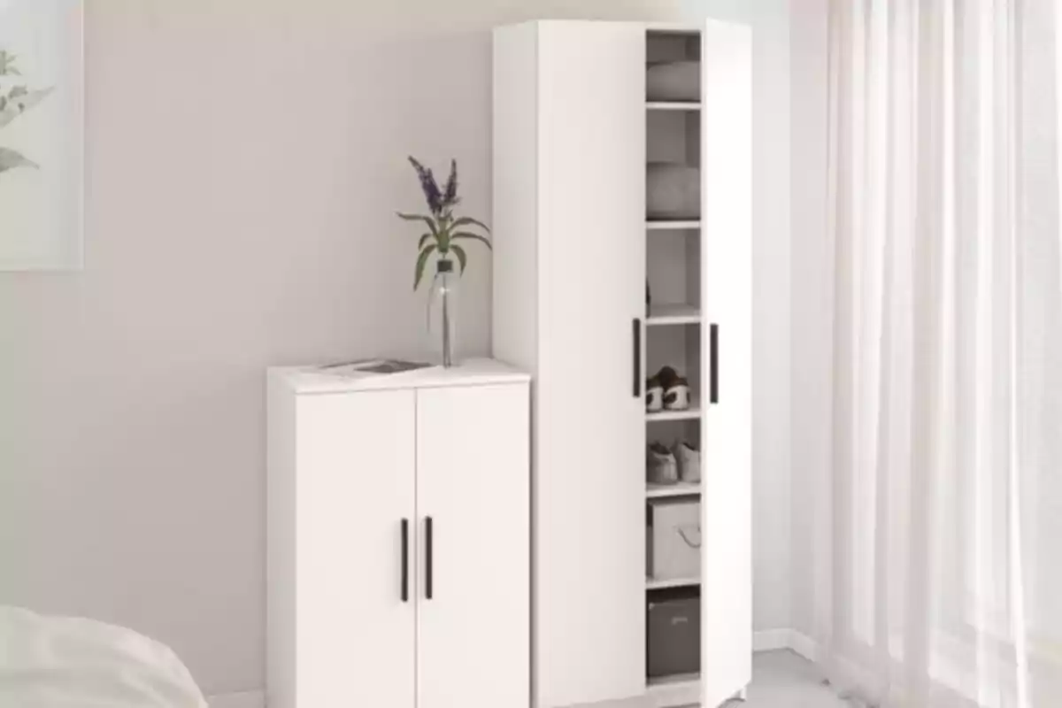 Dos muebles blancos con puertas y tiradores negros junto a una pared clara, uno de ellos con una puerta entreabierta mostrando estantes con ropa y cajas, y el otro con un florero encima Dos muebles blancos con puertas y tiradores negros junto a una pared clara, uno de ellos con una puerta entreabierta mostrando estantes con ropa y cajas, y el otro con un florero encima