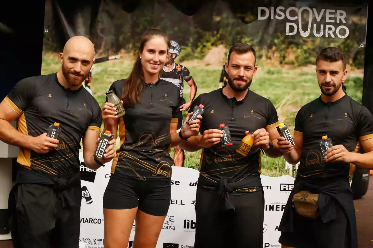 Cuatro personas con ropa deportiva negra posan sonrientes mostrando bebidas energéticas frente a un cartel de evento deportivo al aire libre