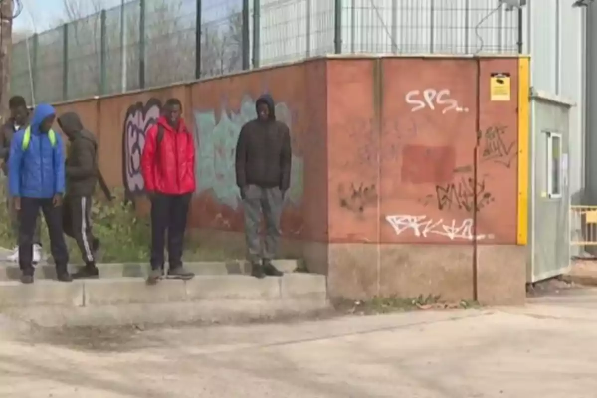 Varios jóvenes con chaquetas y mochilas están de pie junto a una pared con grafitis en una zona urbana