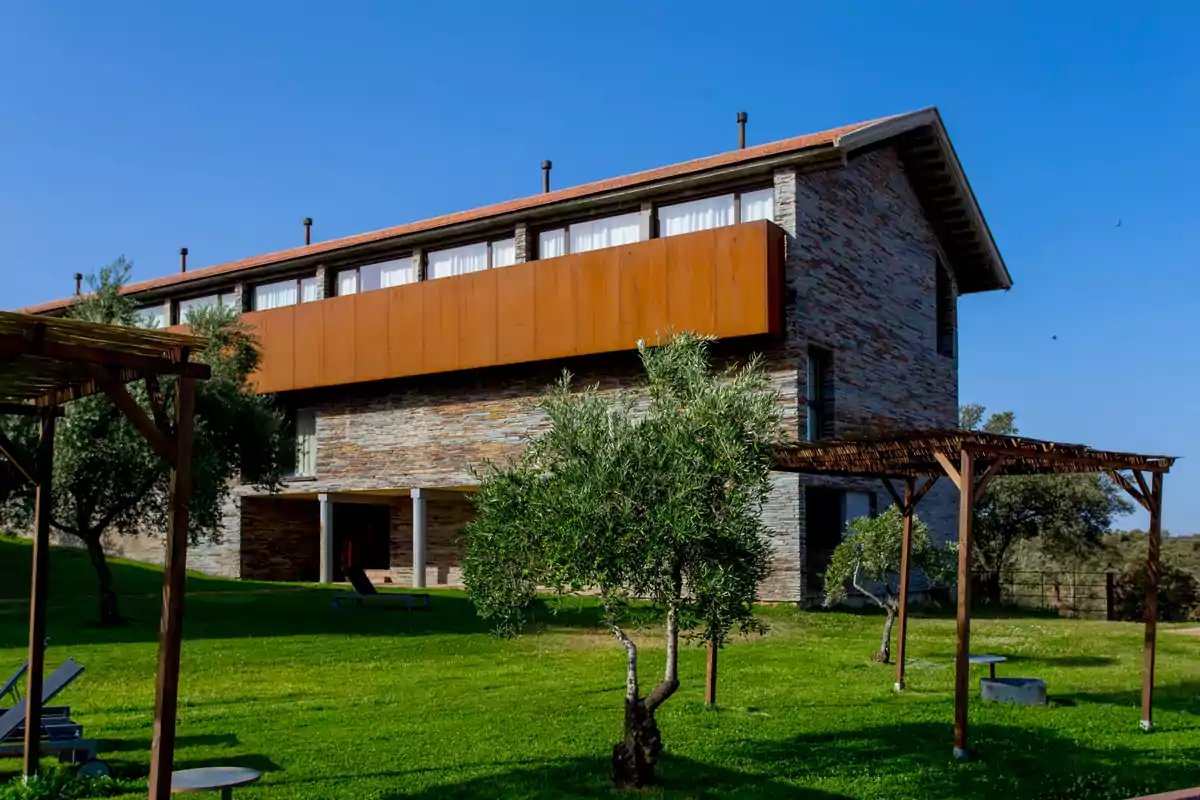 Edificio moderno de piedra y madera con ventanales grandes rodeado de césped verde y árboles bajo un cielo despejado Edificio moderno de piedra y madera con ventanales grandes rodeado de césped verde y árboles bajo un cielo despejado