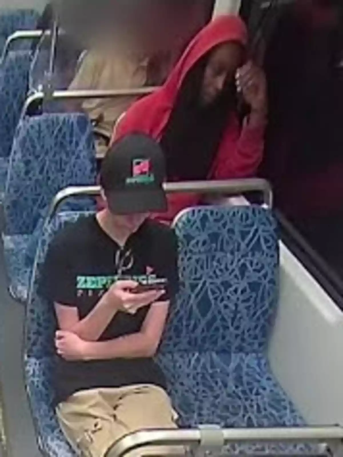 Dos personas sentadas en asientos azules de transporte público, una con sudadera roja y la otra con gorra negra mirando un teléfono móvil.