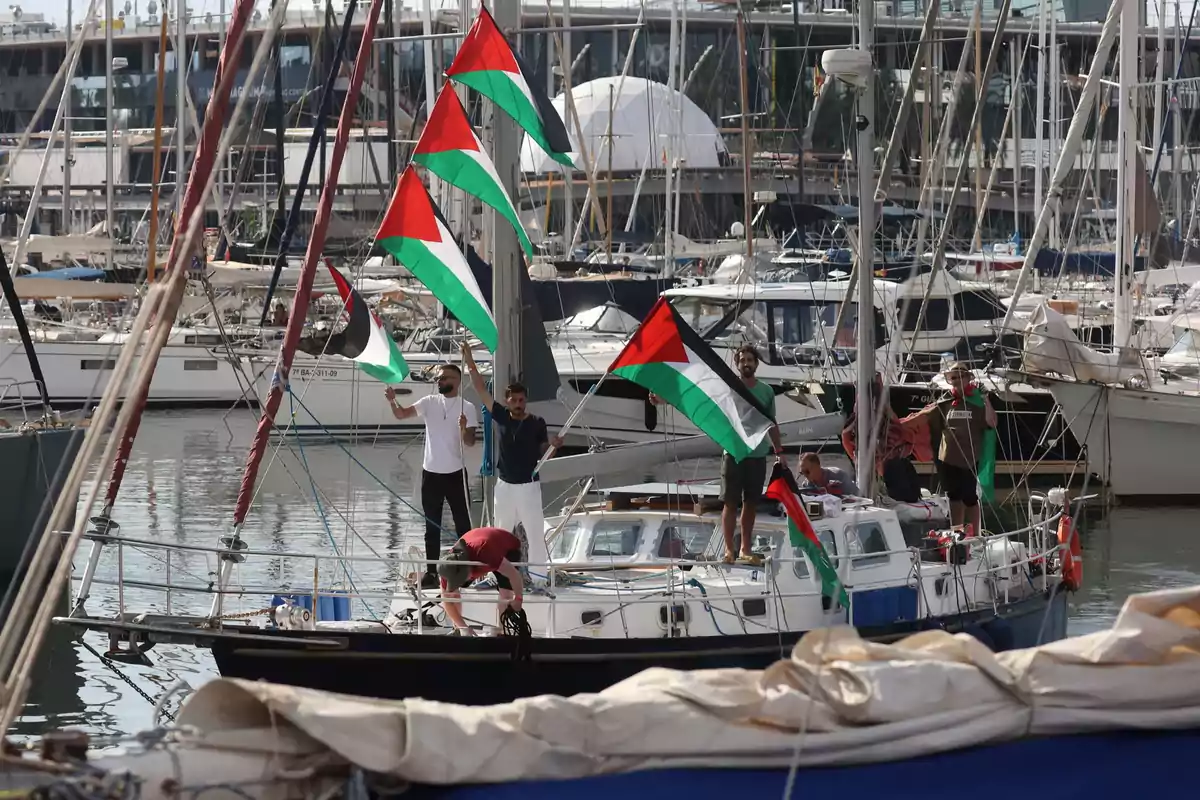 Varias personas en un barco en un puerto sostienen banderas palestinas rodeados de otros barcos y yates Varias personas en un barco en un puerto sostienen banderas palestinas rodeados de otros barcos y yates