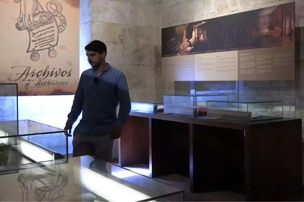 Un hombre camina dentro de una sala de exhibición con vitrinas de cristal y paneles informativos en las paredes, el ambiente es de museo o archivo histórico.
