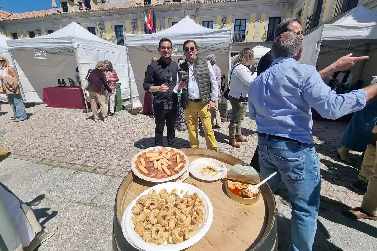 Personas disfrutando de una feria gastronómica al aire libre con platos de comida sobre una mesa de barril y puestos de fondo
