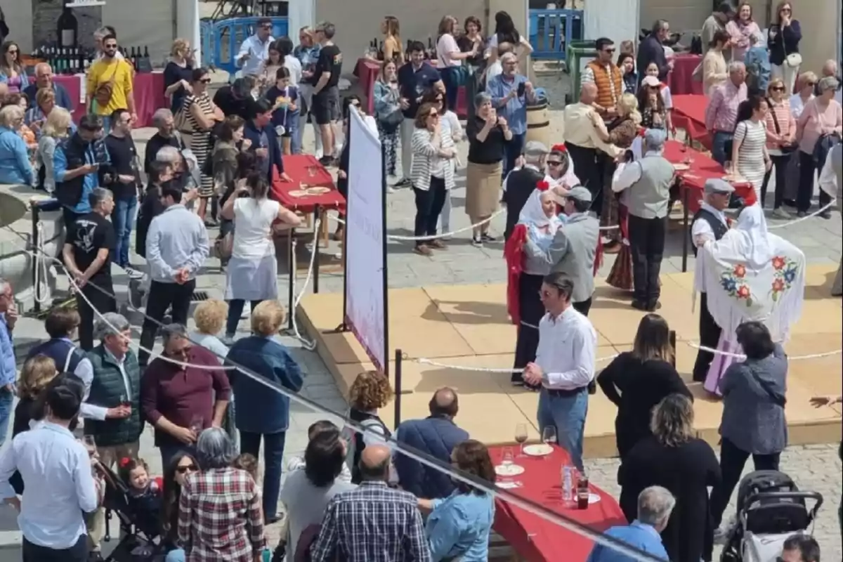 Personas reunidas en una plaza durante una fiesta popular con mesas rojas, comida, bebida y parejas bailando con trajes tradicionales