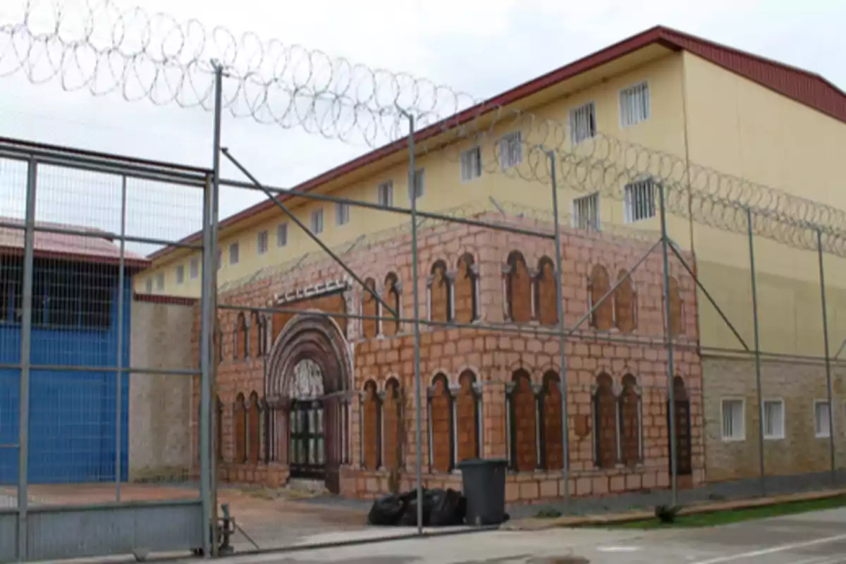 Edificio amarillo con ventanas enrejadas y alambre de púas en la parte superior, detrás de una cerca metálica, con una pared pintada simulando una fachada de piedra Edificio amarillo con ventanas enrejadas y alambre de púas en la parte superior, detrás de una cerca metálica, con una pared pintada simulando una fachada de piedra