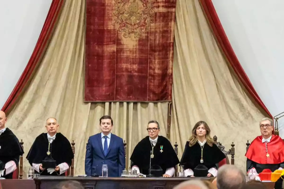 Ceremonia académica con varias personas sentadas en una mesa principal, algunas con toga y medallas, frente a un gran tapiz rojo y beige