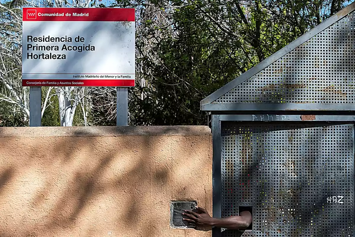 Un letrero de la Residencia de Primera Acogida Hortaleza en Madrid junto a una pared y una puerta metálica por la que sale una mano Un letrero de la Residencia de Primera Acogida Hortaleza en Madrid junto a una pared y una puerta metálica por la que sale una mano