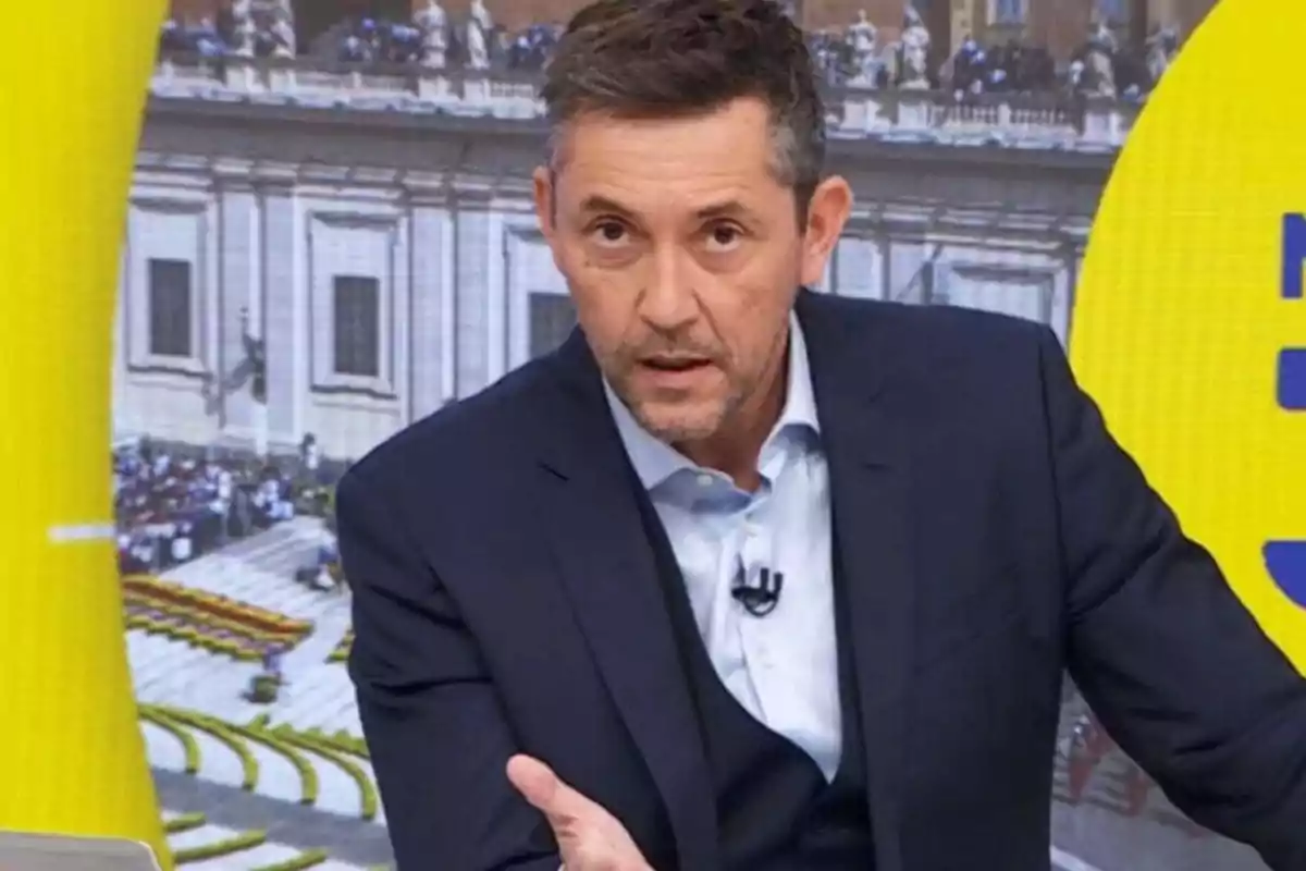 Hombre de cabello corto y barba recortada vestido con traje azul oscuro hablando en un set de televisión con fondo de una plaza y elementos amarillos Hombre de cabello corto y barba recortada vestido con traje azul oscuro hablando en un set de televisión con fondo de una plaza y elementos amarillos