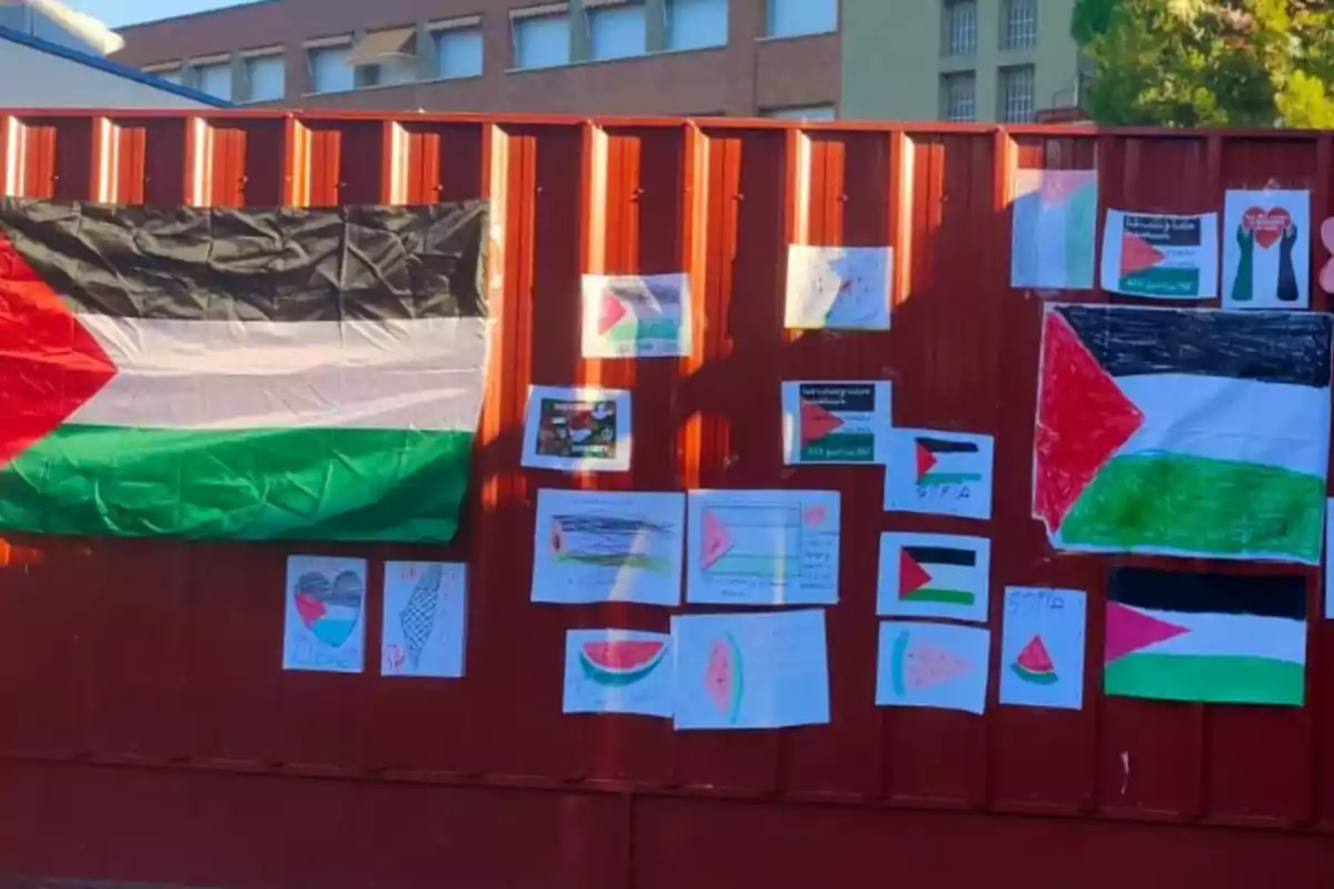 Una cerca de color rojo con varias banderas de Palestina y dibujos infantiles pegados, algunos con sandías y corazones, en un entorno urbano.