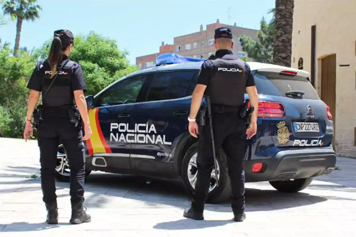 Dos agentes de la Policía Nacional de España de espaldas junto a un coche patrulla en una zona urbana y soleada