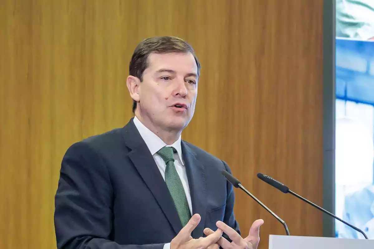 Hombre de traje oscuro y corbata verde hablando frente a dos micrófonos con fondo de madera Hombre de traje oscuro y corbata verde hablando frente a dos micrófonos con fondo de madera
