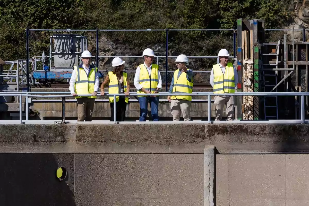 Cinco personas con chalecos reflectantes y cascos blancos observan una obra de construcción desde una barandilla