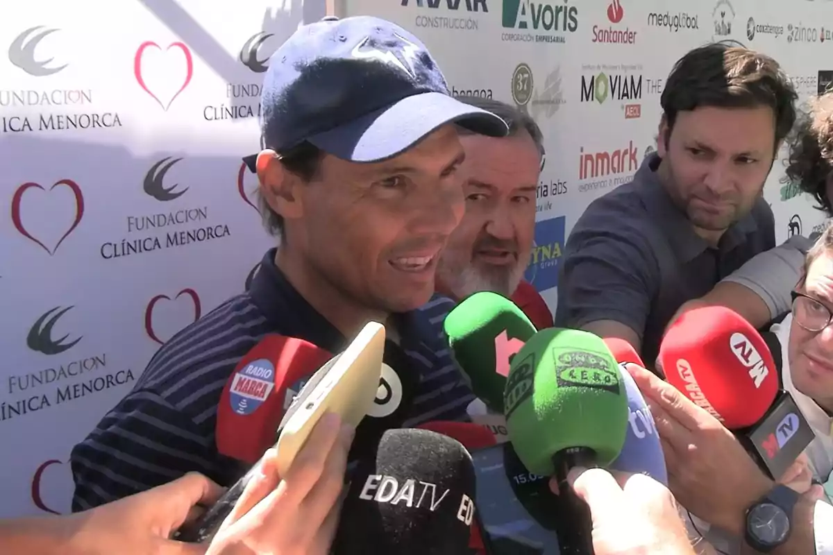 Un hombre con gorra azul da una entrevista rodeado de varios micrófonos de diferentes medios de comunicación frente a un fondo con logotipos de patrocinadores. Un hombre con gorra azul da una entrevista rodeado de varios micrófonos de diferentes medios de comunicación frente a un fondo con logotipos de patrocinadores.