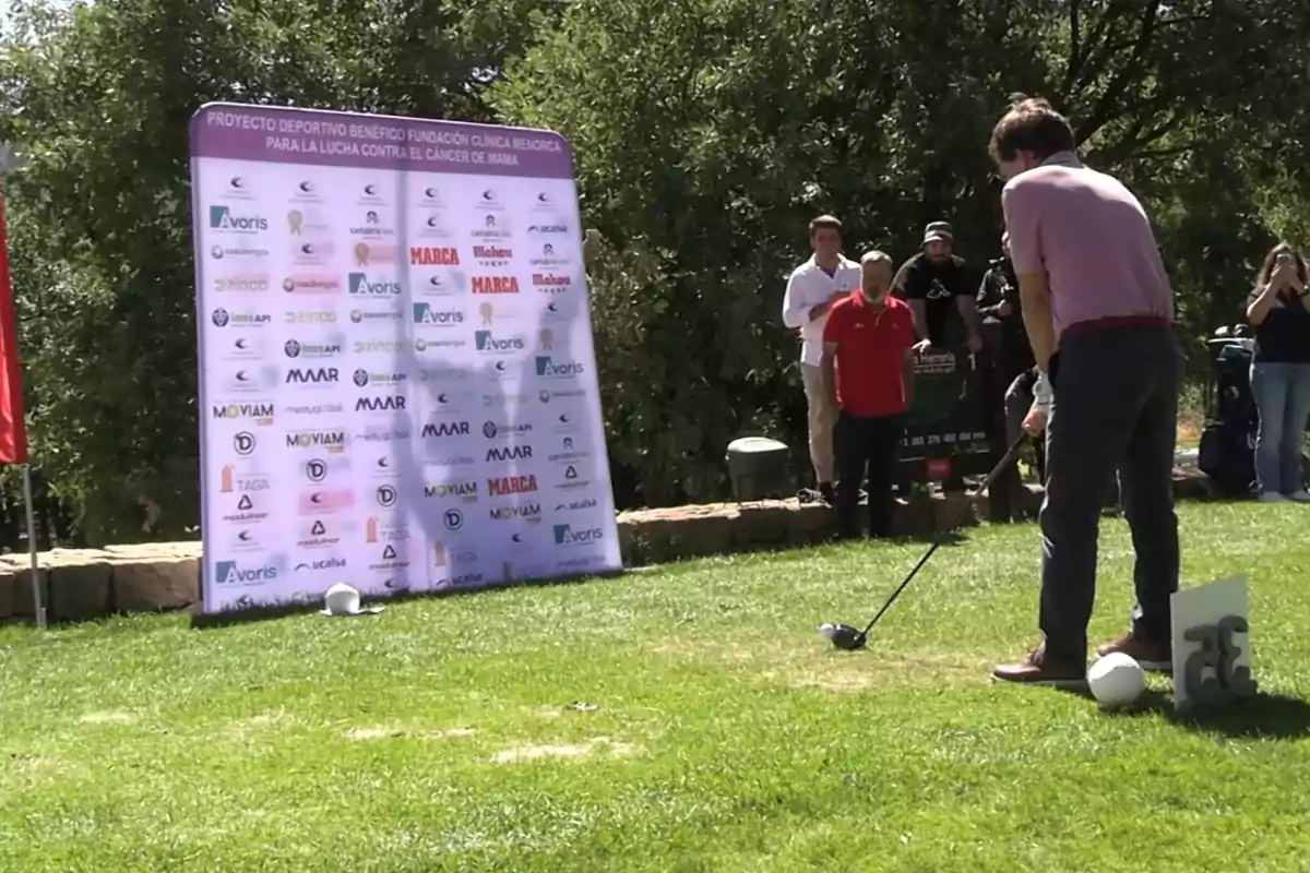 Un hombre se prepara para golpear una pelota de golf mientras varias personas observan y un cartel con logotipos de patrocinadores está al fondo en un evento al aire libre. Un hombre se prepara para golpear una pelota de golf mientras varias personas observan y un cartel con logotipos de patrocinadores está al fondo en un evento al aire libre.