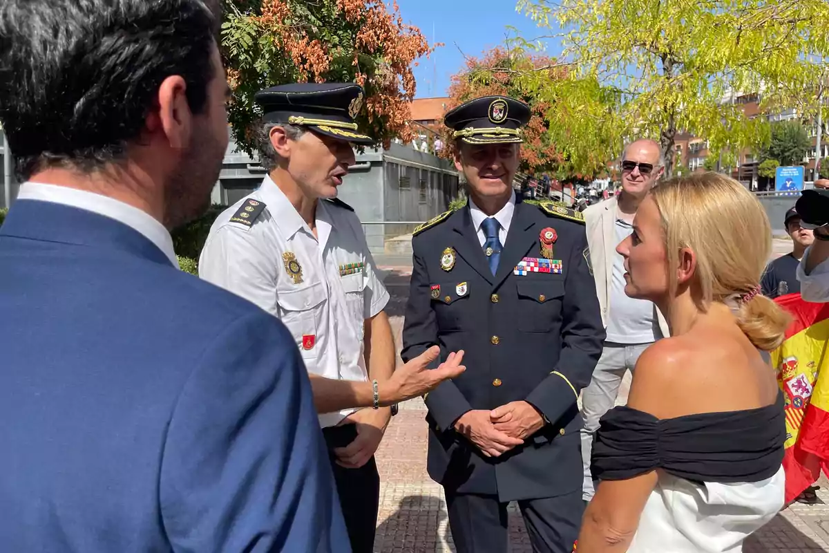 Varias personas conversan al aire libre, entre ellas dos hombres con uniformes de policía y una mujer rubia vestida de blanco y negro, mientras al fondo se observa una bandera de España y árboles con hojas verdes y naranjas. Varias personas conversan al aire libre, entre ellas dos hombres con uniformes de policía y una mujer rubia vestida de blanco y negro, mientras al fondo se observa una bandera de España y árboles con hojas verdes y naranjas.