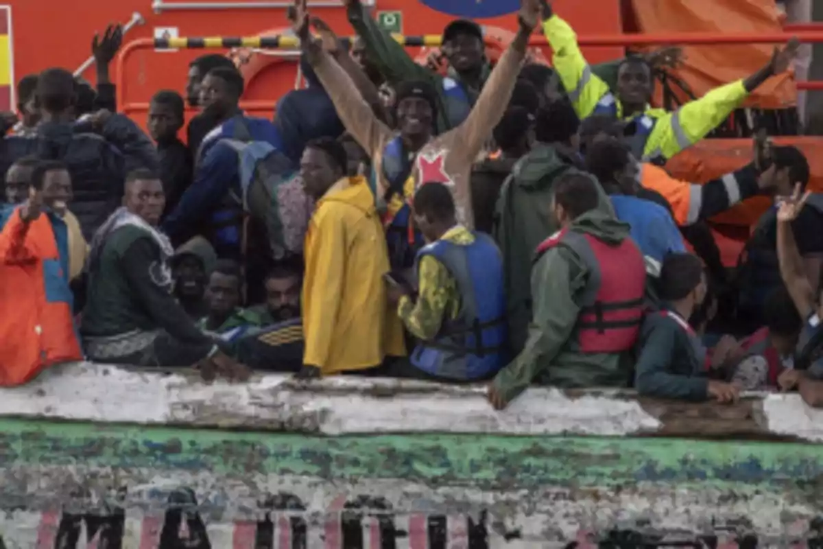 Un grupo numeroso de personas con chalecos salvavidas viaja en una embarcación de madera, algunas levantan los brazos y otras miran hacia la cámara, al fondo se observa una estructura naranja de un barco de rescate. Un grupo numeroso de personas con chalecos salvavidas viaja en una embarcación de madera, algunas levantan los brazos y otras miran hacia la cámara, al fondo se observa una estructura naranja de un barco de rescate.