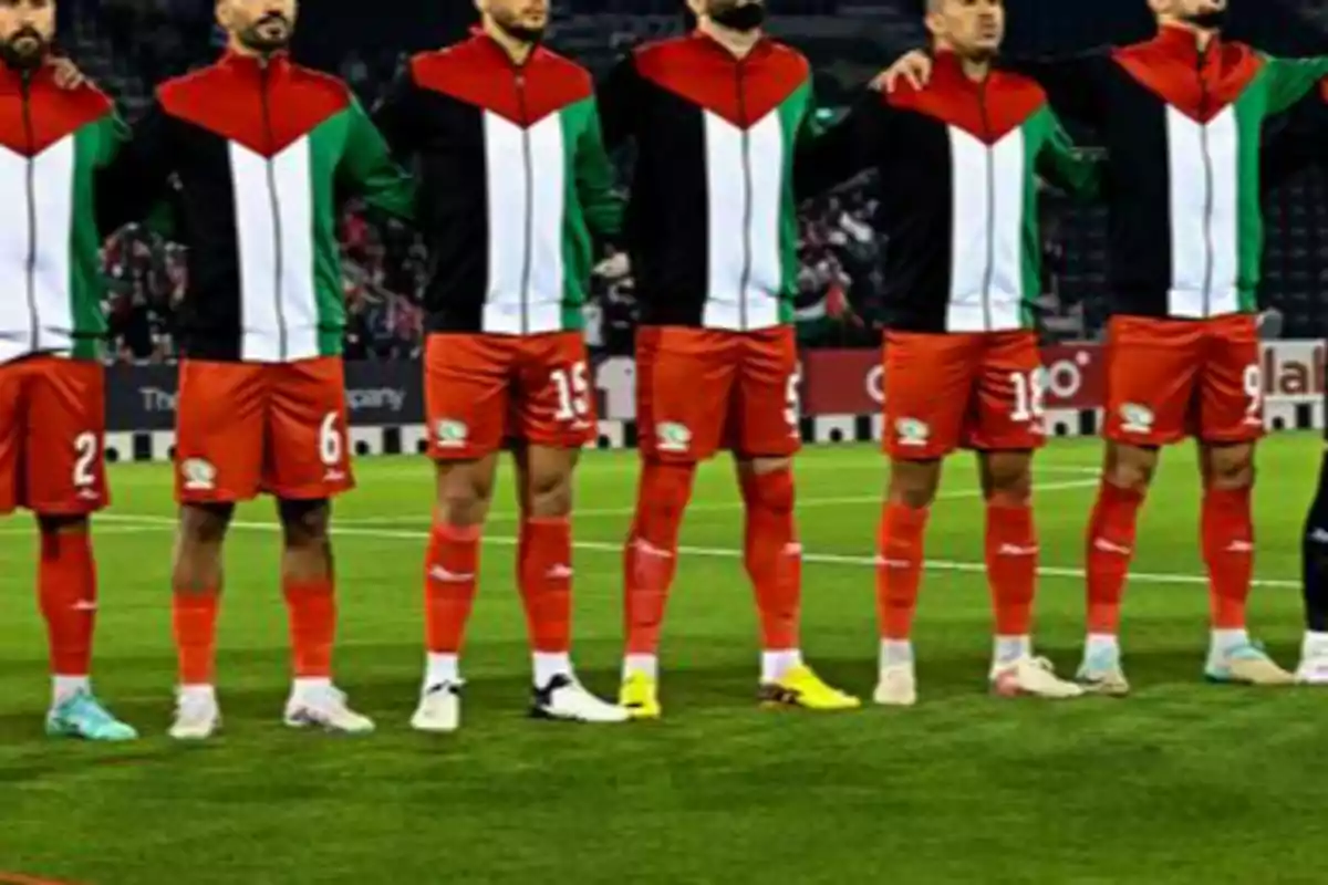 Jugadores de fútbol con uniformes rojos y chaquetas con los colores de la bandera de Palestina están formados en el campo antes de un partido Jugadores de fútbol con uniformes rojos y chaquetas con los colores de la bandera de Palestina están formados en el campo antes de un partido