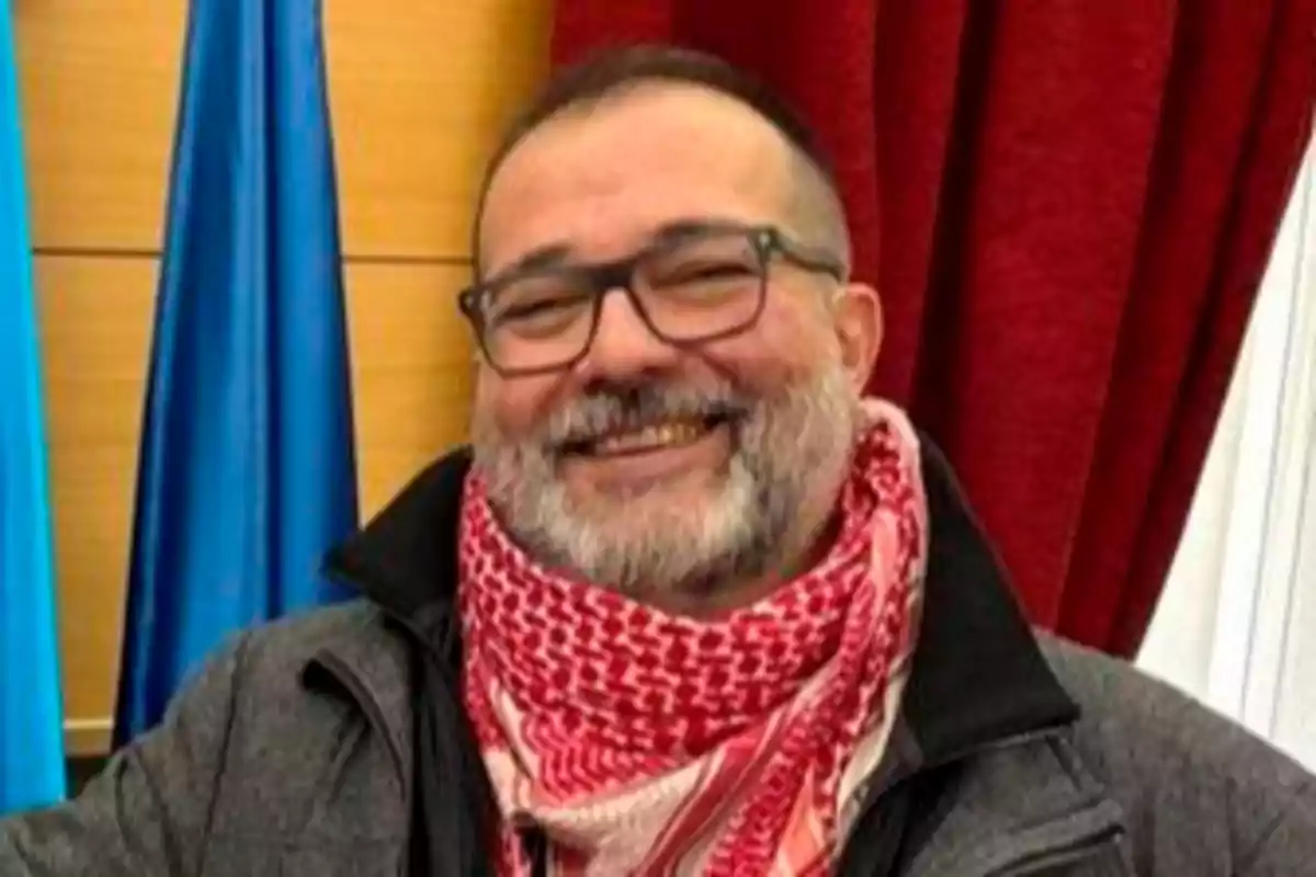 Hombre de barba y gafas sonriendo, lleva una bufanda roja y blanca y un abrigo gris, con cortinas rojas y banderas azules de fondo