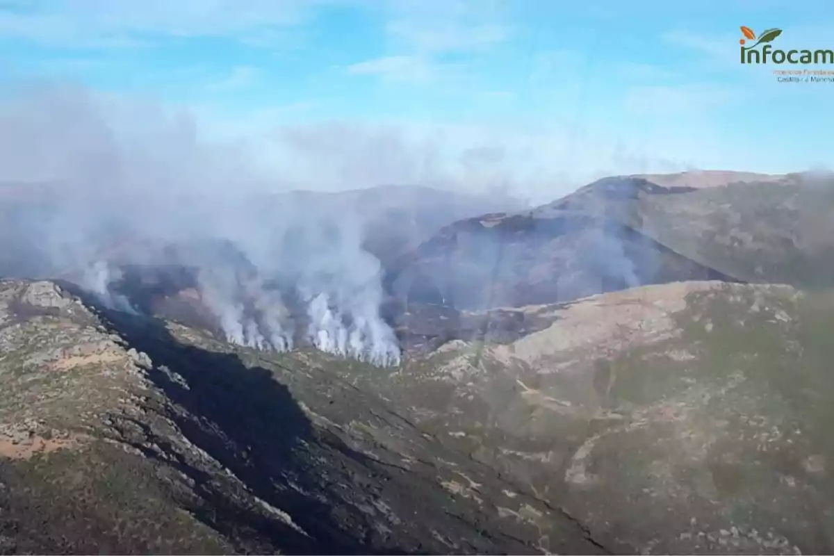 Incendio forestal en una zona montañosa con columnas de humo elevándose entre las colinas bajo un cielo azul