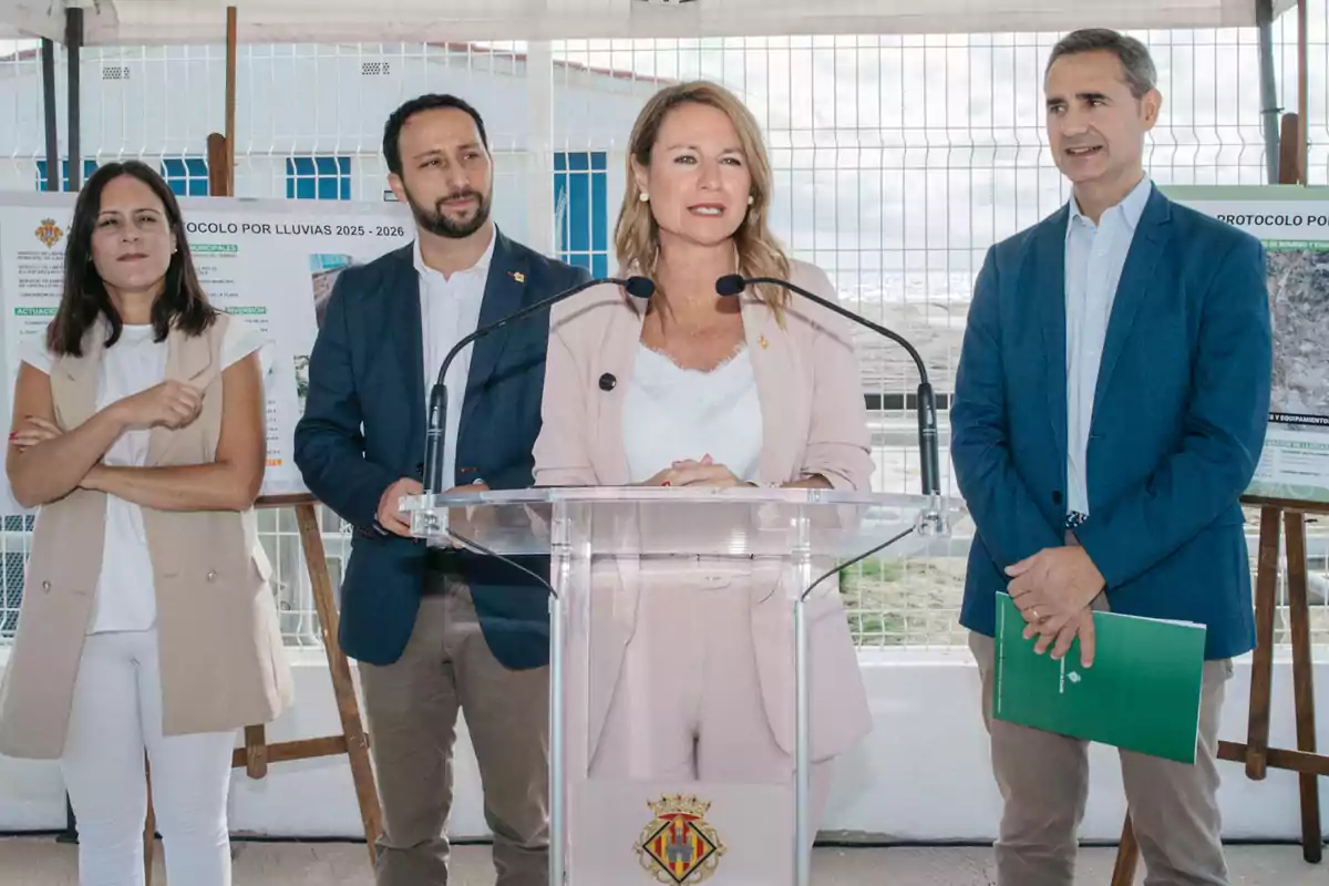 Cuatro personas vestidas de manera formal están de pie, una mujer al centro habla en un atril transparente con micrófonos, mientras las otras tres personas la acompañan, al fondo hay carteles informativos y una reja blanca.