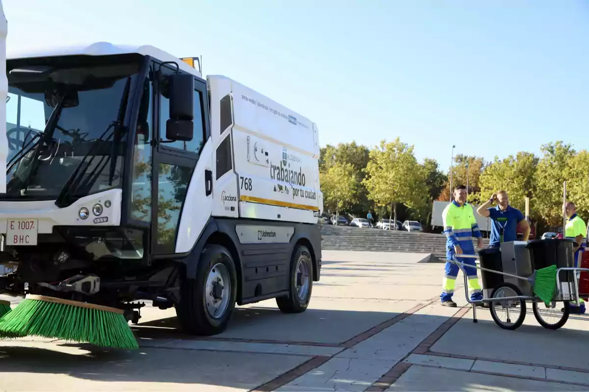 Camión barredora de limpieza urbana junto a tres trabajadores con uniforme de alta visibilidad y un carrito de basura en una zona peatonal con árboles al fondo