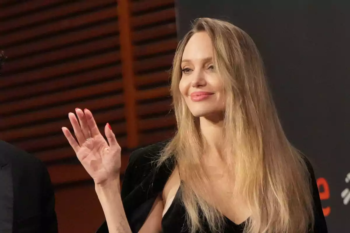 Mujer de cabello rubio largo sonriendo y saludando con la mano derecha en un evento formal