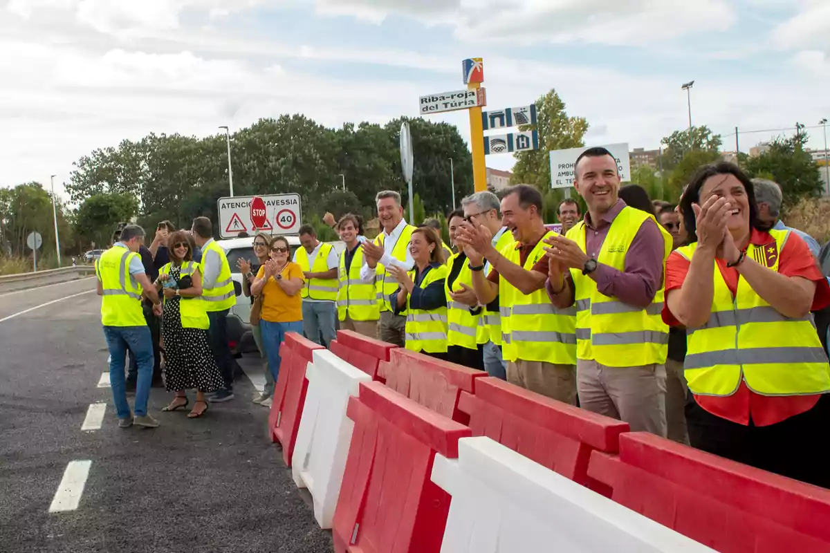 Un grupo de personas con chalecos reflectantes aplaude y sonríe junto a una carretera recién asfaltada con barreras de seguridad rojas y blancas en primer plano