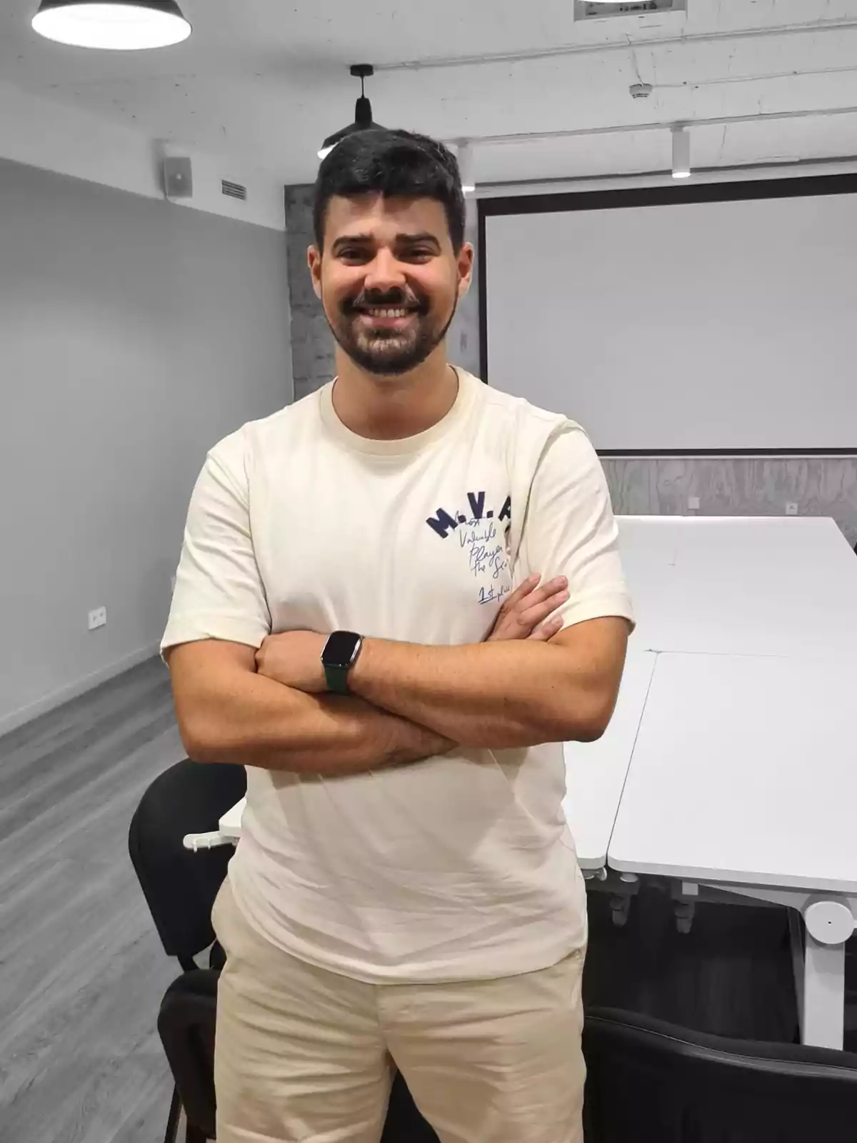 Hombre joven de cabello oscuro y barba sonríe con los brazos cruzados en una sala de reuniones moderna con mesas y sillas blancas